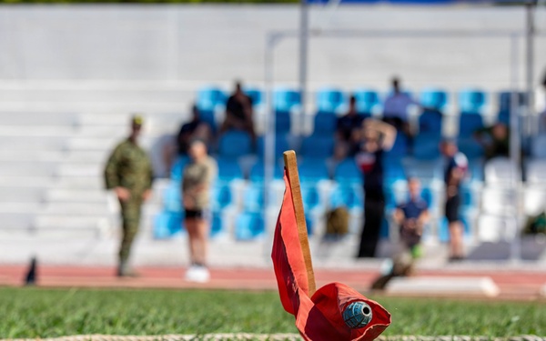 The image shows a close-up of a red marker flag with a wooden stick planted in green grass at what appears to be a military or athletic competition venue. Two rope lines are visible on the grass, likely marking boundaries or lanes for a field event. In the blurred background, individuals in military uniforms and civilian clothing are visible near stadium seating with blue chairs.