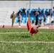 The image shows a close-up of a red marker flag with a wooden stick planted in green grass at what appears to be a military or athletic competition venue. Two rope lines are visible on the grass, likely marking boundaries or lanes for a field event. In the blurred background, individuals in military uniforms and civilian clothing are visible near stadium seating with blue chairs.
