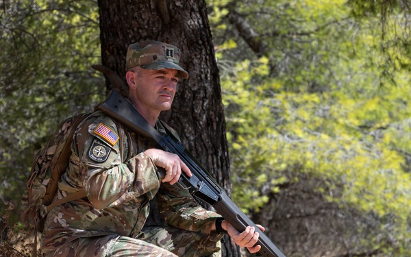 A male soldier in U.S. Army MultiCam (OCP) camouflage uniform is crouching against a tree in a wooded, rocky outdoor environment. He is holding a rifle and wearing a patrol cap with what appears to be a captain's rank insignia. Patches on his left shoulder include a U.S. flag and a unit patch.