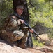 A male soldier in U.S. Army MultiCam (OCP) camouflage uniform is crouching against a tree in a wooded, rocky outdoor environment. He is holding a rifle and wearing a patrol cap with what appears to be a captain's rank insignia. Patches on his left shoulder include a U.S. flag and a unit patch.