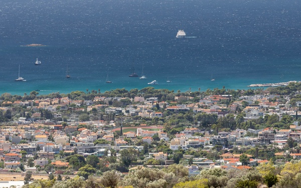 An elevated aerial view of a Mediterranean coastal town with white and terracotta-roofed buildings nestled between a hillside and a deep blue sea. The foreground features a dry, rocky hillside covered with olive trees and scrub vegetation, with a dirt trail where a small group of hikers is visible. Several sailboats and yachts are anchored in the turquoise coastal waters.