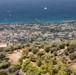 An elevated aerial view of a Mediterranean coastal town with white and terracotta-roofed buildings nestled between a hillside and a deep blue sea. The foreground features a dry, rocky hillside covered with olive trees and scrub vegetation, with a dirt trail where a small group of hikers is visible. Several sailboats and yachts are anchored in the turquoise coastal waters.