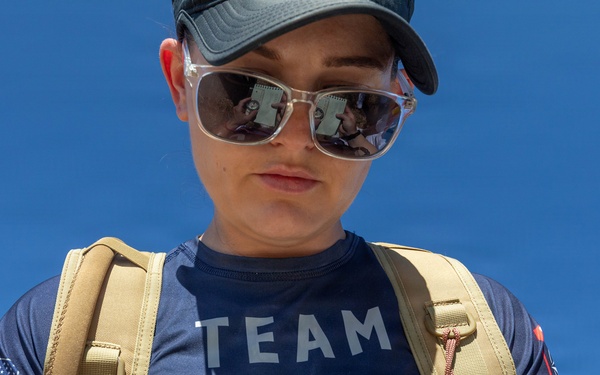 A woman wearing a 'Team USA' navy blue t-shirt and a tan tactical backpack is looking down at a small notebook or notepad she is holding. She is wearing sunglasses and a dark baseball cap, standing outdoors under a clear blue sky. An American flag patch is visible on her left sleeve.