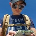 A woman wearing a 'Team USA' navy blue t-shirt and a tan tactical backpack is looking down at a small notebook or notepad she is holding. She is wearing sunglasses and a dark baseball cap, standing outdoors under a clear blue sky. An American flag patch is visible on her left sleeve.