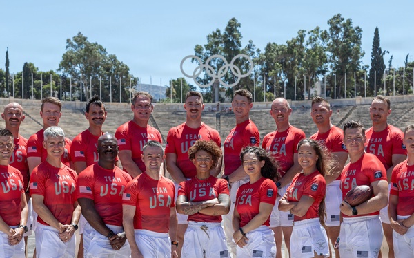 A group of approximately 22 athletes and staff pose for a team photo wearing red 'Team USA' uniforms with white shorts at what appears to be the Panathenaic Stadium in Athens, Greece. The Olympic rings are clearly visible in the background atop the stadium structure. One person is holding an American football, suggesting this is a flag football or American football delegation.