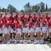 A group of approximately 22 athletes and staff pose for a team photo wearing red 'Team USA' uniforms with white shorts at what appears to be the Panathenaic Stadium in Athens, Greece. The Olympic rings are clearly visible in the background atop the stadium structure. One person is holding an American football, suggesting this is a flag football or American football delegation.