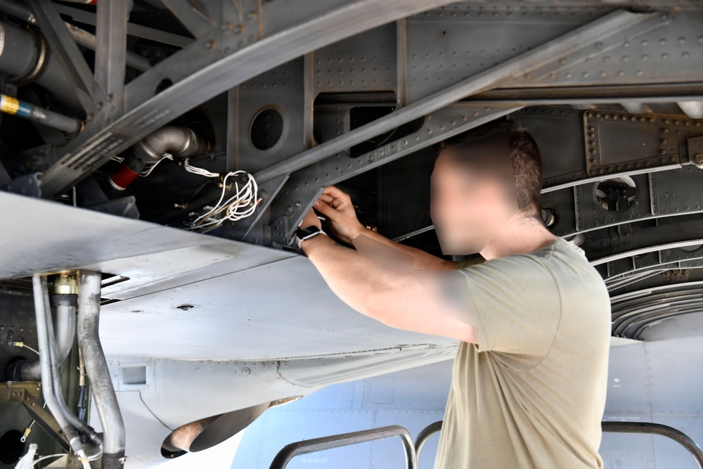 A technician in a olive/tan t-shirt is performing maintenance work on the underside of a military aircraft. The person is reaching upward into an open access panel area, working with wiring or connectors visible in the aircraft's internal structure. The subject's face has been intentionally blurred, suggesting operational security considerations.