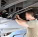 A technician in a olive/tan t-shirt is performing maintenance work on the underside of a military aircraft. The person is reaching upward into an open access panel area, working with wiring or connectors visible in the aircraft's internal structure. The subject's face has been intentionally blurred, suggesting operational security considerations.