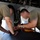 Two maintenance technicians in olive/tan military-style t-shirts are working underneath an aircraft, performing what appears to be electrical or avionics maintenance. They are leaning into an open access panel or landing gear bay area, using test equipment including a multimeter with red and black leads. The faces of both individuals have been intentionally obscured/blurred.