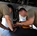 Two maintenance technicians in olive/tan military-style t-shirts are working underneath an aircraft, performing what appears to be electrical or avionics maintenance. They are leaning into an open access panel or landing gear bay area, using test equipment including a multimeter with red and black leads. The faces of both individuals have been intentionally obscured/blurred.