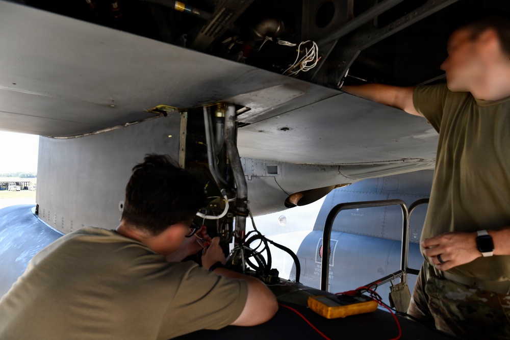 Two military maintenance technicians are performing work on the underside of a large military aircraft, likely in a landing gear bay or weapons bay area. One technician is closely inspecting or repairing hydraulic or electrical components while wearing a headset, while the second technician observes and assists. A yellow multimeter with red and black test leads is visible, indicating electrical diagnostic work is being performed.