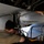 Two military maintenance technicians are performing work on the underside of a large military aircraft, likely in a landing gear bay or weapons bay area. One technician is closely inspecting or repairing hydraulic or electrical components while wearing a headset, while the second technician observes and assists. A yellow multimeter with red and black test leads is visible, indicating electrical diagnostic work is being performed.
