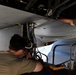 Two military maintenance technicians are performing work on the underside of a large military aircraft, likely in a landing gear bay or weapons bay area. One technician is closely inspecting or repairing hydraulic or electrical components while wearing a headset, while the second technician observes and assists. A yellow multimeter with red and black test leads is visible, indicating electrical diagnostic work is being performed.
