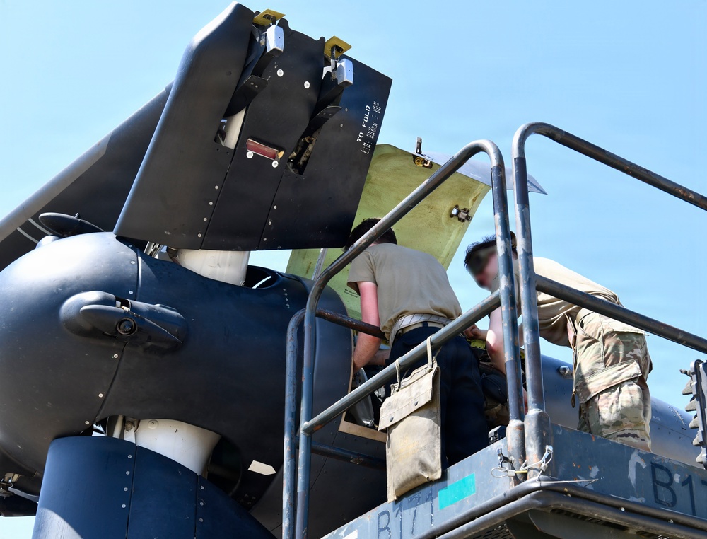 Two maintenance personnel are working on what appears to be a military aircraft, likely performing maintenance or inspection on a folded wing section or engine nacelle area. One individual is wearing a camouflage military uniform while the other is in a tan/beige shirt. They are positioned on an elevated maintenance platform or stand marked with 'CB1' or 'B17' lettering.