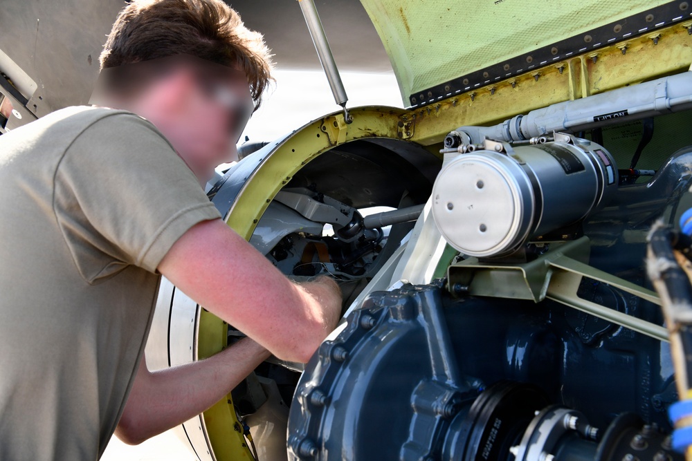 A technician in a tan/olive t-shirt is performing maintenance work on what appears to be a military or large aircraft engine compartment. The individual is leaning into an open access panel, working on internal mechanical components. The face has been intentionally blurred, suggesting this is an official or sensitive military/government image.