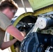 A technician in a tan/olive t-shirt is performing maintenance work on what appears to be a military or large aircraft engine compartment. The individual is leaning into an open access panel, working on internal mechanical components. The face has been intentionally blurred, suggesting this is an official or sensitive military/government image.