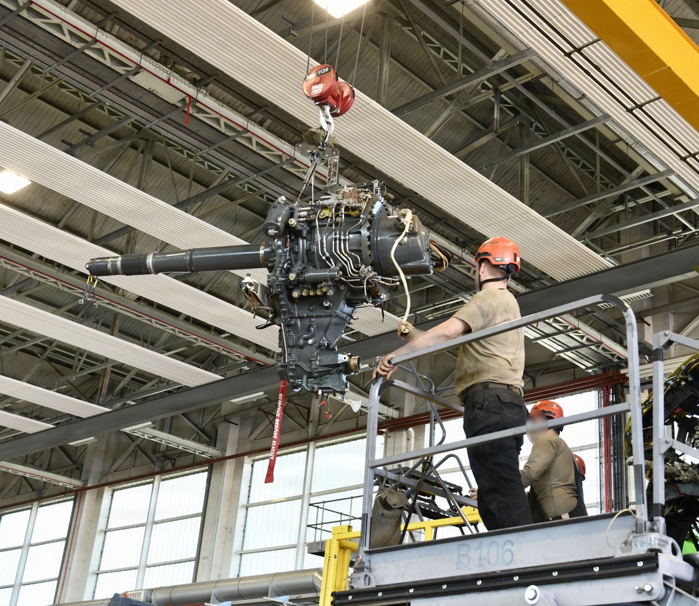 Two workers wearing orange safety helmets are operating on an elevated scissor lift platform labeled 'B106' inside a large industrial hangar or maintenance facility. They are handling a large mechanical assembly — appearing to be a helicopter main rotor gearbox or transmission — suspended by an overhead crane with a red hook block. The setting is a well-lit aerospace or military maintenance bay with a yellow overhead crane rail system visible.
