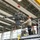 Two workers wearing orange safety helmets are operating on an elevated scissor lift platform labeled 'B106' inside a large industrial hangar or maintenance facility. They are handling a large mechanical assembly — appearing to be a helicopter main rotor gearbox or transmission — suspended by an overhead crane with a red hook block. The setting is a well-lit aerospace or military maintenance bay with a yellow overhead crane rail system visible.