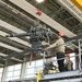Two workers wearing orange safety helmets are operating on an elevated scissor lift platform labeled 'B106' inside a large industrial hangar or maintenance facility. They are handling a large mechanical assembly — appearing to be a helicopter main rotor gearbox or transmission — suspended by an overhead crane with a red hook block. The setting is a well-lit aerospace or military maintenance bay with a yellow overhead crane rail system visible.
