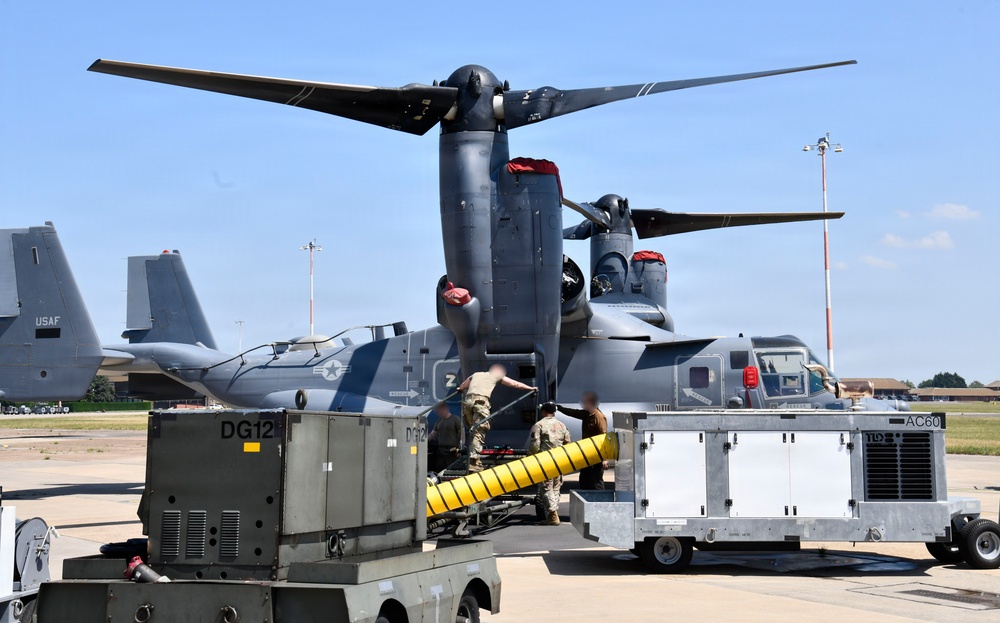 A military tiltrotor aircraft, identifiable as a CV-22 or V-22 Osprey in dark gray livery, is undergoing ground servicing operations on a flight line. Ground support equipment including power units labeled 'DG12' and 'AC60' are positioned alongside the aircraft, with uniformed personnel actively working between them. A USAF aircraft tail is visible in the background, indicating a U.S. Air Force installation.