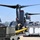A military tiltrotor aircraft, identifiable as a CV-22 or V-22 Osprey in dark gray livery, is undergoing ground servicing operations on a flight line. Ground support equipment including power units labeled 'DG12' and 'AC60' are positioned alongside the aircraft, with uniformed personnel actively working between them. A USAF aircraft tail is visible in the background, indicating a U.S. Air Force installation.