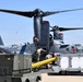 A military tiltrotor aircraft, identifiable as a CV-22 or V-22 Osprey in dark gray livery, is undergoing ground servicing operations on a flight line. Ground support equipment including power units labeled 'DG12' and 'AC60' are positioned alongside the aircraft, with uniformed personnel actively working between them. A USAF aircraft tail is visible in the background, indicating a U.S. Air Force installation.