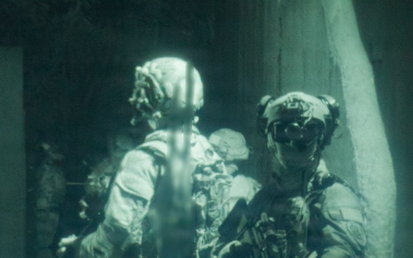 The image shows multiple armed military personnel in full tactical gear positioned near a doorway or entry point of a structure. The photograph has a distinctive green-tinted, grainy quality consistent with night-vision imagery or infrared photography. The soldiers appear to be conducting a building entry or clearing operation.