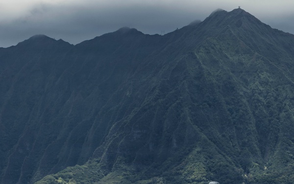 A military C-130 Hercules transport aircraft with tail number '0037' is parked on a tarmac, with a line of personnel boarding or disembarking near the rear of the aircraft. The scene is set against a dramatic backdrop of steep, lush green volcanic mountains consistent with the Ko'olau Range in Hawaii, likely at Marine Corps Base Hawaii (Kaneohe Bay). Residential and commercial structures are visible at the base of the mountains.