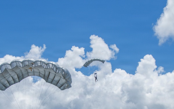 A military parachute training exercise is taking place over a large open field with mountainous terrain in the background. Multiple paratroopers are visible at various altitudes descending with gray/silver ram-air parachutes. The setting appears to be a military airfield or drop zone, likely in Hawaii based on the distinctive mountain ridgeline.