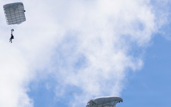 Three parachutists are descending through a partly cloudy blue sky, each suspended beneath a large white square-shaped parachute canopy. The parachutes appear to be military-style ram-air or round canopies in white/light gray coloring. The image is taken from a low angle looking upward, capturing all three jumpers at different altitudes.