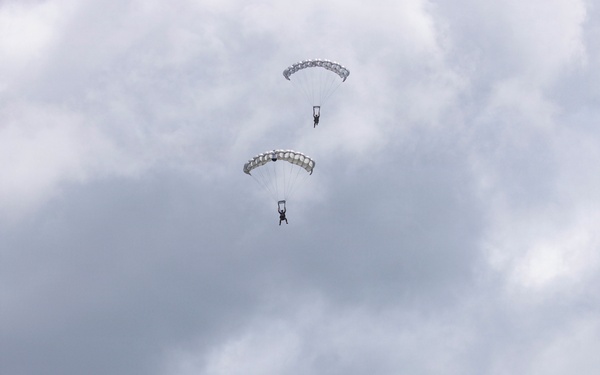Three parachutists are descending in a vertical formation against an overcast, cloudy sky with snow-capped mountain peaks visible at the bottom of the frame. All three parachutes are fully deployed and appear to be white/silver ram-air or round canopy type. The image captures a military or sport skydiving operation in a mountainous environment.