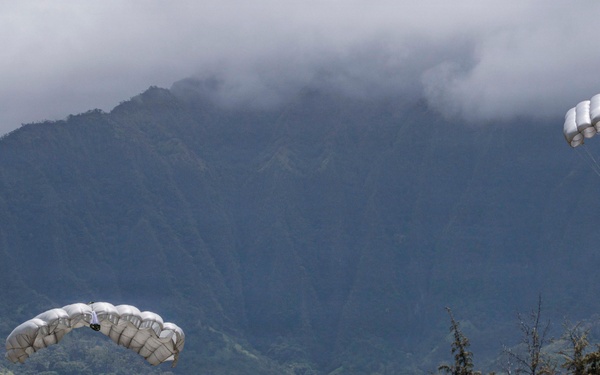 A military parachute training exercise is underway on a grassy drop zone, with multiple paratroopers landing or having already landed. Two parachutists are still airborne with deployed white/silver ram-air canopies, while several others are on the ground managing collapsed chutes. The scene is set against a dramatic backdrop of steep, cloud-shrouded green mountains consistent with Hawaii's Ko'olau Range.