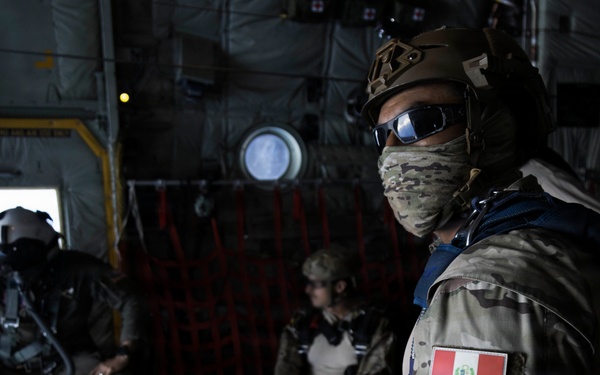 Three military personnel are inside what appears to be a military transport aircraft, likely a C-130 or similar cargo plane. The foreground soldier wears Peruvian military insignia (Peruvian flag patch and a medical/unit patch) along with a tactical helmet, goggles, and a camouflage face covering. The scene suggests a pre-jump or airborne operation preparation.
