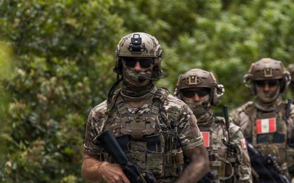 Four heavily armed and equipped special operations soldiers are moving in a patrol formation along a path surrounded by dense green vegetation. At least three of the soldiers display Peruvian flag patches on their left shoulders, identifying them as members of the Peruvian military. The lead soldier carries an assault rifle and is equipped with advanced tactical gear including a helmet-mounted night vision device mount and communications equipment.