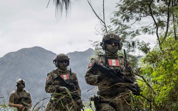 Three armed soldiers in camouflage uniforms are conducting a patrol along a jungle trail in a mountainous, heavily vegetated environment. At least two of the soldiers display Peruvian flag patches on their left shoulders, identifying them as members of the Peruvian military. The soldiers are equipped with tactical gear, helmets, sunglasses, and assault rifles.