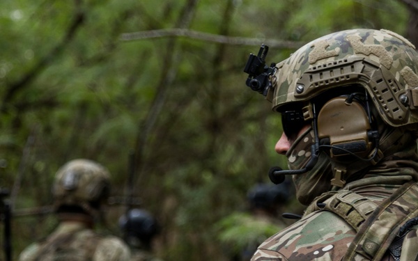 The image shows a group of military personnel in a forested environment, equipped with full tactical gear. The foreground soldier is in sharp focus wearing multicam camouflage, a ballistic helmet with mounted device, communications headset, face covering, plate carrier vest, and a backpack. Additional soldiers are visible in the background, out of focus, similarly equipped.