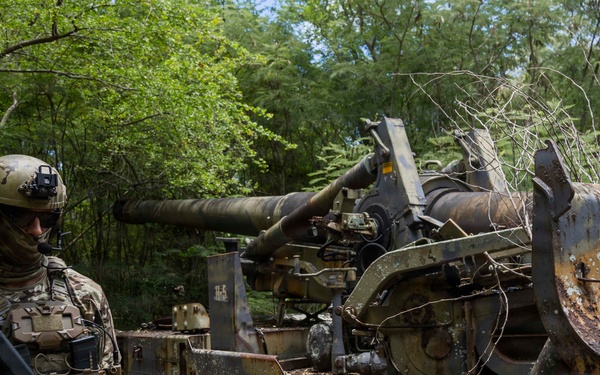 A heavily equipped soldier in multicam camouflage stands beside a heavily rusted and deteriorated tracked military vehicle, likely an abandoned self-propelled artillery piece, in a dense forested area. The vehicle shows significant corrosion, peeling paint, and vegetation growth indicating long-term abandonment. The soldier appears to be inspecting or documenting the derelict equipment.