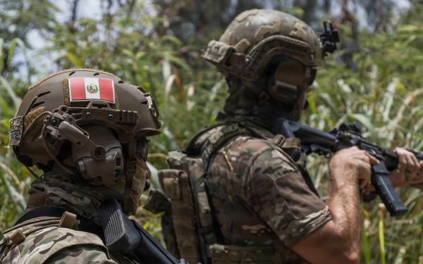 Two armed military personnel in multicam camouflage uniforms are positioned in dense vegetation, appearing to conduct a patrol or tactical operation. The soldier in the foreground displays Peruvian flag patches on both the helmet and left shoulder, identifying them as members of the Peruvian armed forces. Both soldiers are carrying assault rifles and wearing full tactical gear including helmets and plate carriers.
