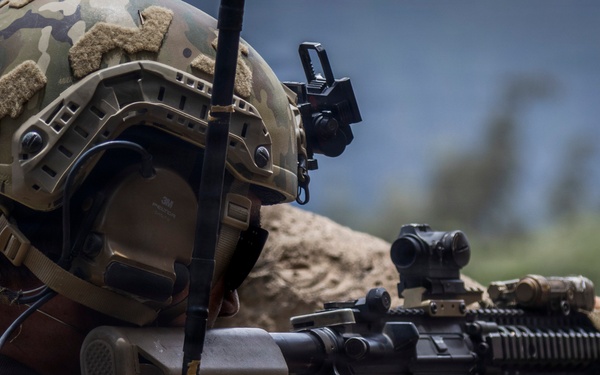 A heavily equipped soldier in multicam camouflage is positioned behind a rock or stone barrier, aiming a rifle with an optic sight. The soldier is wearing advanced tactical gear including a ballistic helmet with mounted accessories, communications headset, and body armor. The background shows a blurred outdoor environment suggesting a mountainous or arid terrain.