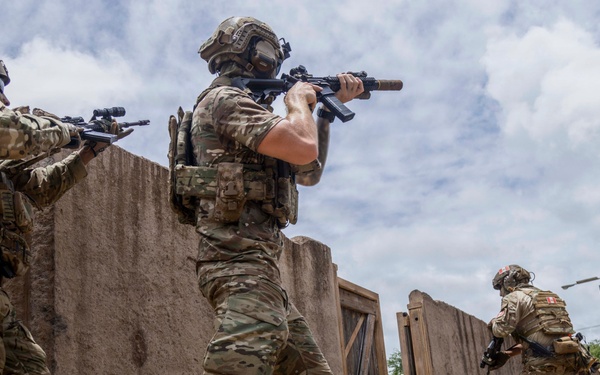 The image shows a group of four armed military or special operations personnel conducting what appears to be a tactical training exercise in an urban warfare simulation environment. The soldiers are moving in formation through a compound with adobe-style walls and structures, with weapons raised in a ready position. The setting appears to be a purpose-built Military Operations on Urban Terrain (MOUT) training facility.