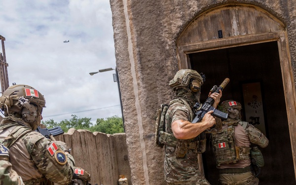 A group of heavily armed military or special operations personnel in multicam camouflage are conducting a building entry/clearing exercise or operation. Multiple operators are visible wearing helmets with night vision mounts, body armor, and carrying rifles, with at least one preparing to breach a doorway of a mud-brick structure. Patches visible on their uniforms appear to include Peruvian flag insignia, suggesting these may be Peruvian special forces.