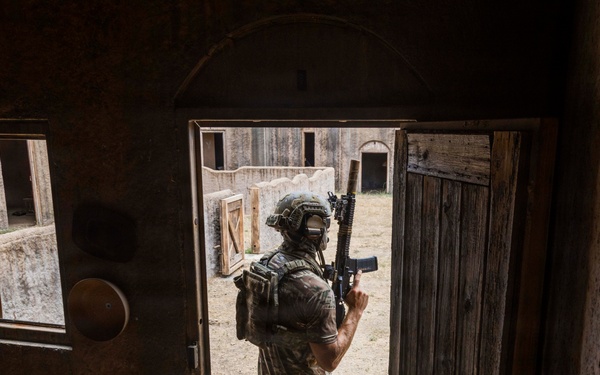 A soldier in full tactical gear is seen from behind, standing in a doorway of a rustic building and scanning an open courtyard area. The individual is equipped with a rifle, combat helmet, and tactical vest, suggesting a military training exercise or operational scenario. The environment appears to be a mock village or training facility based on the constructed wooden structures and sandy terrain.