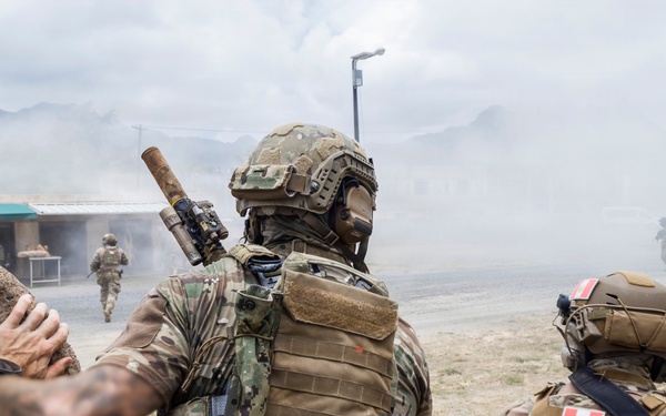 Two armed soldiers in tactical gear observe a smoke-filled training area from behind a large rock formation. One soldier displays a Peruvian flag patch on their shoulder, suggesting a joint or multinational military exercise. Additional personnel are visible moving through the smoke in the background.