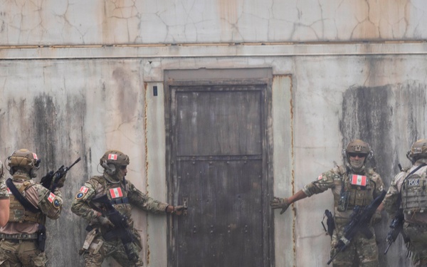 A group of military personnel in full combat gear are conducting a tactical building entry/clearing exercise at what appears to be a training facility. Multiple soldiers are stacked near a door on a weathered concrete structure, executing a coordinated breach maneuver. Shoulder patches visible on the uniforms appear to include Canadian flag insignia, suggesting Canadian Armed Forces participation.