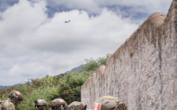 A group of armed military or special operations personnel are taking cover behind a vehicle and a concrete wall, appearing to be conducting a tactical operation or training exercise. The soldiers are wearing full combat gear including tactical helmets, body armor, and carrying assault rifles. A military aircraft is visible in the sky in the background.