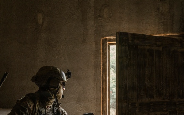 Two armed military or special operations personnel are conducting a room-clearing operation inside a dimly lit structure. The lead operator is aiming a suppressed rifle toward an open wooden door, while a second operator follows behind. The scene depicts tactical close-quarters combat training or an active operation.