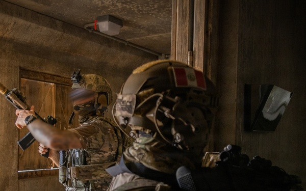 Two heavily equipped military or special operations personnel are conducting a room-clearing exercise inside a building structure. Both operators are wearing multicam camouflage uniforms and tactical helmets, with Peruvian flag patches visible on their gear. The scene appears to be a close-quarters battle (CQB) training scenario.