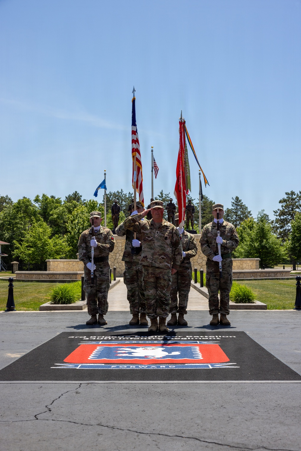 A formal U.S. Army military ceremony is taking place outdoors at what appears to be a memorial or parade ground. A central figure in Army Combat Uniform (ACU) is rendering a salute while flanked by color guard soldiers holding rifles at attention. The ground displays a unit insignia mat reading '3rd Brigade Engineer Battalion, 34th Infantry Division' with the word 'FORWARD' and a red and blue unit crest.