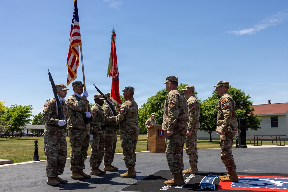 A formal U.S. Army military ceremony is taking place outdoors on a paved platform, likely a change of command or unit activation/deactivation ceremony. Soldiers in Army Combat Uniforms (ACU/OCP pattern) are participating, with a color guard presenting the U.S. flag and unit guidons. The ceremony appears to be conducted at a military installation on a clear, sunny day.