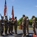 A formal U.S. Army military ceremony is taking place outdoors on a paved platform, likely a change of command or unit activation/deactivation ceremony. Soldiers in Army Combat Uniforms (ACU/OCP pattern) are participating, with a color guard presenting the U.S. flag and unit guidons. The ceremony appears to be conducted at a military installation on a clear, sunny day.